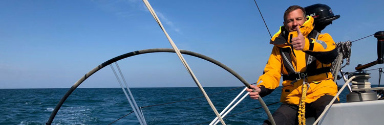 Foto a bordo de un velero. Con una persona en abrigo naranja teniendo el timón y mirando hacia el objetivo con el pulgar levantado. 