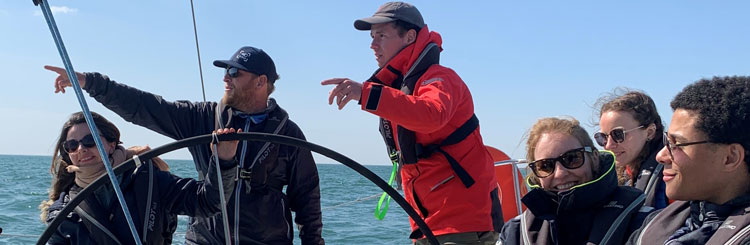 Enfoque desde un barco sobre un grupo de personas sentadas cerca del timón. Dos personas de pie indicando el horizonte y una persona tiene el timón.